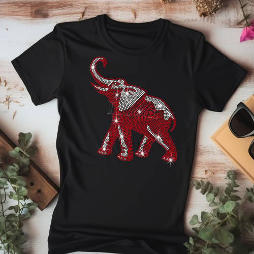 A black t-shirt with a red and silver rhinestone elephant design on the front, displayed on a wooden surface surrounded by green foliage and sunglasses.